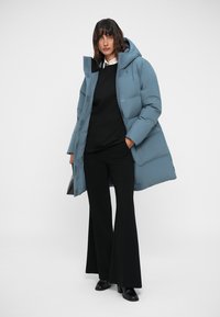 Ljust blå pufferjacka med huva, med en strömlinjeformad design, vadderad textur och frontdragkedja. Bärs över svarta byxor och tröja.