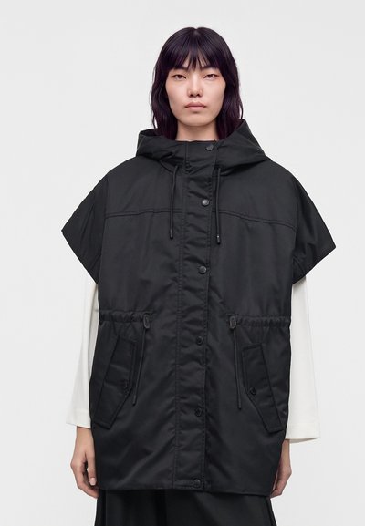 Човек, облечен с черен безръкавен суичър с качулка, копчета и връзки, върху бяла блуза с дълги ръкави, стоящ пред сив фон.