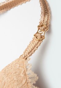 Beżowy koronkowy biustonosz z misternymi detalami, z teksturowanym paskiem i zapięciem w kolorze złotym. Zbliżenie podkreśla wzór.