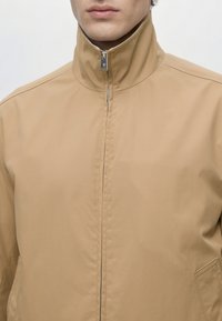 Homme en veste beige zippée avec col haut, ne montrant que le bas du visage et le haut du torse sur un fond uni.