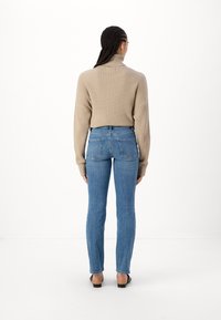 Beige ribbed turtleneck neulepaita, jossa on pudotetut olkapäät, yhdistettynä tiukkoihin sinisiin farkkuihin ja mustiin kenkiin, nähtynä takaa yksinkertaista taustaa vasten.