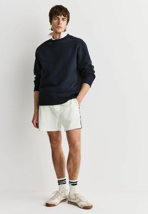Junger Mann, der einen marineblauen Sweatshirt, weißes Polohemd, weiße Shorts, gestreifte Socken und beige Sneakers vor weißem Hintergrund trägt.