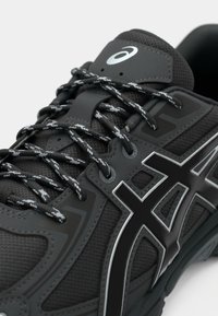 Gros plan d'une chaussure de course Asics noire avec logo réfléchissant, lacets noirs et gris à motif, et languette rembourrée sur fond blanc.