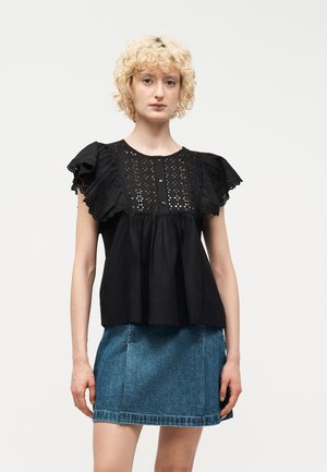 Νεαρό άτομο με κοντά σγουρά ξανθά μαλλιά, που φοράει μαύρο μπλουζάκι με βολάν και λεπτομέρειες με τρύπες, καθώς και μπλε τζιν φούστα, στέκεται μπροστά σε λευκό φόντο.