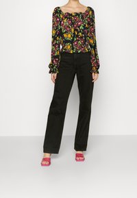 Bloemrijke blouse met lange mouwen in zwart met roze, gele en groene patronen; gecombineerd met hoge zwarte rechte jeans en roze sandalen.