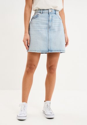 Frau trägt einen hellblauen Jeansrock, ein weißes T-Shirt und weiße High-Top-Sneaker, steht vor einfachem weißen Hintergrund.
