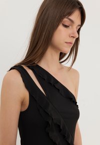 Crna bluzica s jednim ramenom s volanima duž izreza. Izrađena od glatkog materijala s prilagođenim krojem. Jednostavna i elegantna konstrukcija.