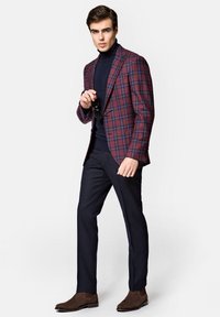 Wzorzysta marynarka w kratę w kolorze czerwonym i granatowym, ciemny golf, dopasowane spodnie oraz brązowe buty Chelsea; mężczyzna trzyma okulary przeciwsłoneczne w prawej ręce.