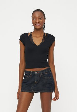 Femme souriante aux cheveux tressés portant un haut noir à col en V avec détail en dentelle et une mini-jupe en jean foncé, sur un fond blanc uni.