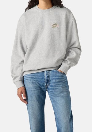 Osoba w jasnoszarej bluzie z niewielkim kwiatowym logo Levi's i jasnoniebieskich dżinsach, z jedną ręką w kieszeni, na jednolitym tle.