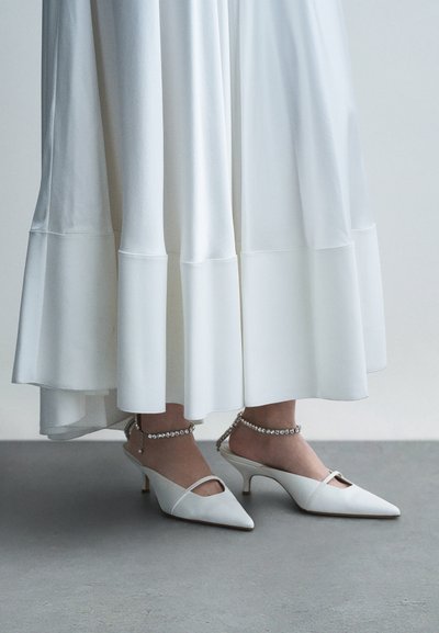 Białe szpilki z wysokim, spiczastym noskiem, otwartymi bokami i paskiem wokół kostki ozdobionym kryształkami, zestawione z lekką, białą spódnicą.