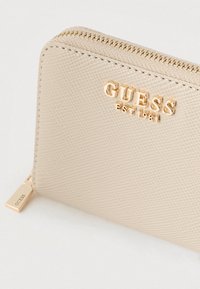 Portafoglio beige con finitura testurizzata, logo in rilievo dorato e chiusura con zip. Presenta un dettaglio in metallo color oro.