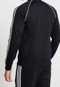 Svart atletisk jacka med vita tre-randsaccent vid axlarna och ärmarna. Tillverkad av slät tyg med en kroppsnära design.