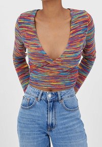 Persoon in een veelkleurige, langarmige gebreide crop top met een diepe V-hals en hoge taille lichtblauwe jeans met knoopsluiting.