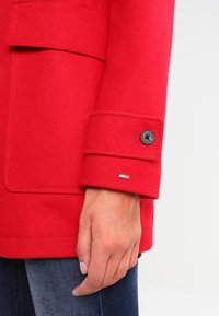 Röd filtrock med knäppt manschett, med en svart knapp och en liten metalllogotyp som accent. Synlig hand och denim under.