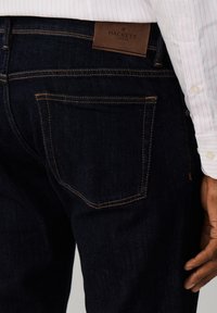 Detailný záber na tmavomodré rifle s oranžovým prešitím a koženou nášivkou Hackett London, nosené s ružovou pruhovanou košeľou a viditeľnou rukou.