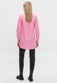 Long-sleeve pink shirt with a straight cut, buttoned cuffs, and a back pleat, paired with black slim-fit pants and chunky black boots.