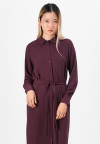 Maroon button-up dress with a collar and long sleeves, featuring a waist tie and textured fabric. Buttons run down the front.