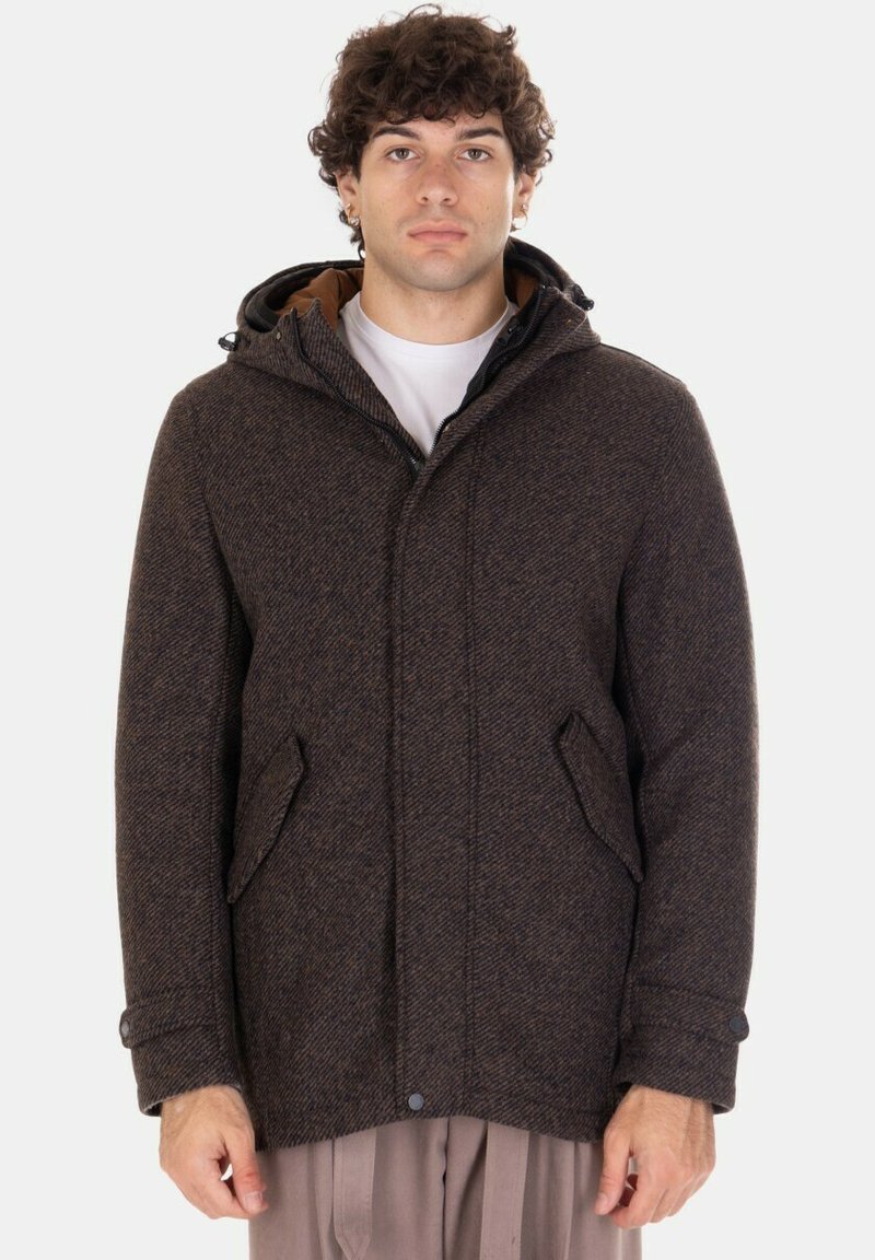 Giovane uomo che indossa un cappotto di lana con cappuccio color marrone scuro, con tasche a patta e polsini con bottoni, in piedi davanti a uno sfondo chiaro e uniforme.