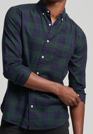 Mand iført en mørkegrøn og navy blå ternet skjorte med opkørte ærmer, justerer et ærme med hånden.