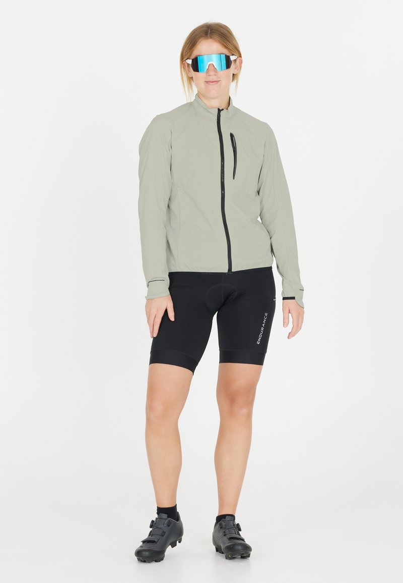 Lys grå cykeljakke med høj krave, sorte cykelshorts og sorte cykelsko. Har store spejlede blå solbriller.