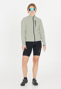 Lys grå cykeljakke med høj krave, sorte cykelshorts og sorte cykelsko. Har store spejlede blå solbriller.