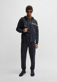Svart hoodie med guld accenter, i kombination med matchande joggingbyxor och mörka sneakers. Texturen ser mjuk ut; designen är atletisk och modern.