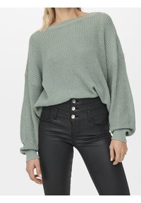 Femme portant un pull ample en tricot vert sauge avec des manches bouffantes, rentré dans un pantalon noir taille haute à quatre boutons argentés.