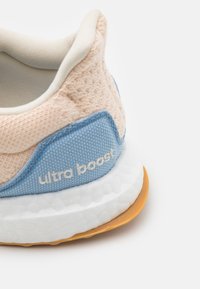 Gros plan sur le talon d'une chaussure de sport beige et bleue avec un tissu texturé, une semelle blanche rembourrée, une semelle extérieure en caoutchouc naturel et le texte "ultra boost" sur une partie bleue.