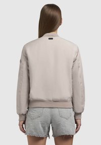 Heledane beez värvi bomber-jakk, millel on soonikkoesäär ja -kätised, tekstuuriga kangas ning väike logo seljal, kantud hallide denimsärkide kohal.