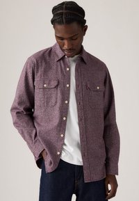 Long-sleeve shirt in a textured burgundy fabric, featuring two chest pockets, cream buttons, and a subtle pattern. Worn over a white t-shirt.