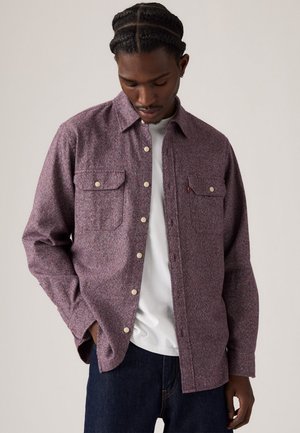 Camisa de manga larga en una tela texturizada color burdeos, con dos bolsillos en el pecho, botones crema y un patrón sutil. Lleva puesta una camiseta blanca debajo.