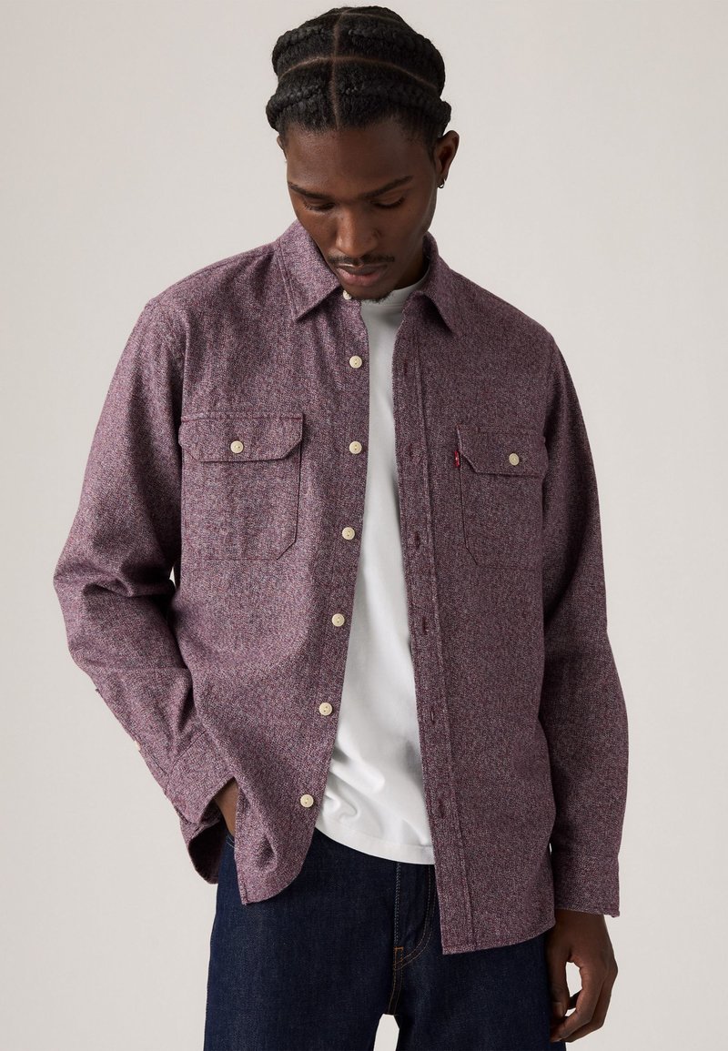 Long-sleeve shirt in a textured burgundy fabric, featuring two chest pockets, cream buttons, and a subtle pattern. Worn over a white t-shirt.
