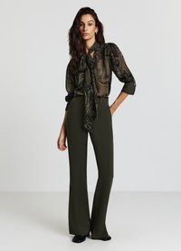 Blusa nera trasparente con intricato motivo paisley, legata al collo, abbinata a pantaloni verde oliva a vita alta. Elegante vestibilità e caduta.