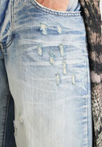 Ljusblå slitna denimjeans med fransade detaljer och en ljusare tvättstruktur. Synlig ficka vid höfterna utan framträdande hårdvara.
