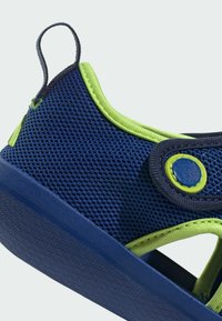 Sininen verkkomateriaali kenkä, jossa on limenvihreitä yksityiskohtia, tekstiurheilupohja ja pyöreä nappisuljenta. Sisältää vetoketjun helpompaa pukemista varten.