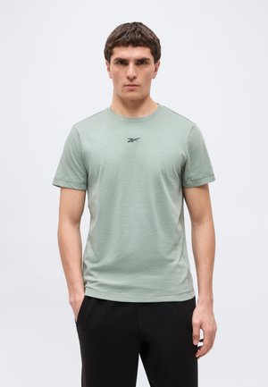 Mladi moški stoji pokončno, oblečen v svetlo zeleno Reebok majico in črne hlače, z rokami delno v žepih.