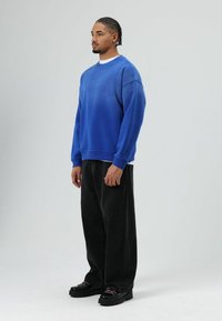 Sweat-shirt bleu avec un design en dégradé, surface texturée, poignets côtelés, associé à un pantalon noir ample et des chaussures noires épaisses.