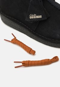 Botte en suede noire avec une surface texturée et un détail de lacets contrastants en beige à côté. La botte présente une semelle cousue et une étiquette de marque.