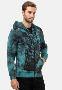 Tealfarbene Kapuzenjacke mit Reißverschluss und Batikeffekt, ausgestattet mit Grafiken, Vorder- Taschen und gerippten Bündchen.