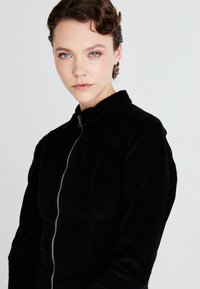 Svart manchesterjacka med hög krage och framzipper, med texturerat tyg och figursydd design, visad mot en neutral bakgrund.