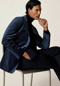 Námornícky sametový blejzer s klapkovým golierom a jedným gombíkom, skombinovaný s čiernym rolákom; model sedí na kovovej stoličke.