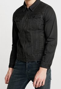 Giacca di jeans nera con zip frontale, due tasche sul petto e colletto. Presenta un design aderente e un tessuto testurizzato.