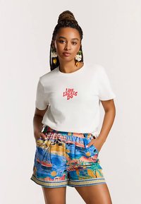 Fehér pamut póló "Like Locals Do" piros felirattal. Színes rövidnadrág tengerparti jelenettel, hajókkal és házakkal, gumis derékrésszel, oldalzsebekkel.