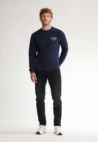Felpa navy con logo "PTRL", collo a giro, maniche lunghe, polsini a coste, abbinata a jeans neri e scarpe da ginnastica bianche. Texture liscia.