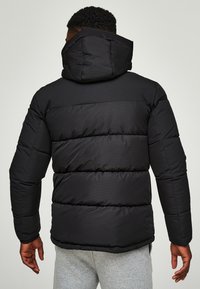 Černá péřová bunda s kapucí, se prošívanými částmi, hladkou texturou a elastickými manžetami. Viditelné švy a volný střih.