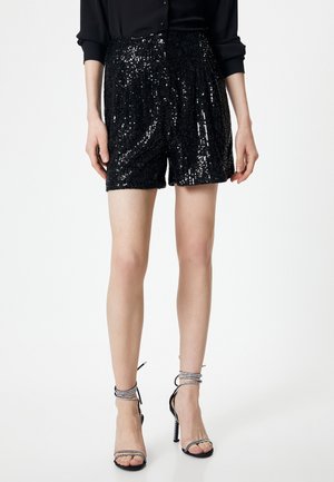 Pantalones cortos de lentejuelas negras con un diseño plisado, que cuentan con una cinturilla ancha. Combinados con sandalias de tacón alto negras con tiras.