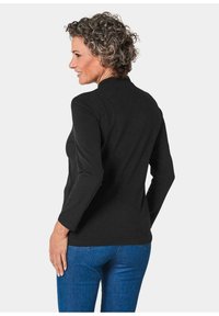 Zwarte top met lange mouwen en hoge hals, strakke pasvorm en gladde textuur, gecombineerd met blauwe jeans. Beeld van achter, met een eenvoudige silhouette.