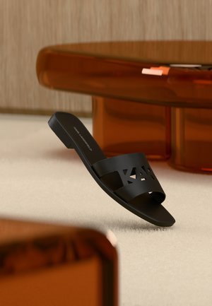 Sandalo nero con suola piatta e marchio ritagliato su un tappeto chiaro in una stanza con mobili traslucidi di colore ambra.