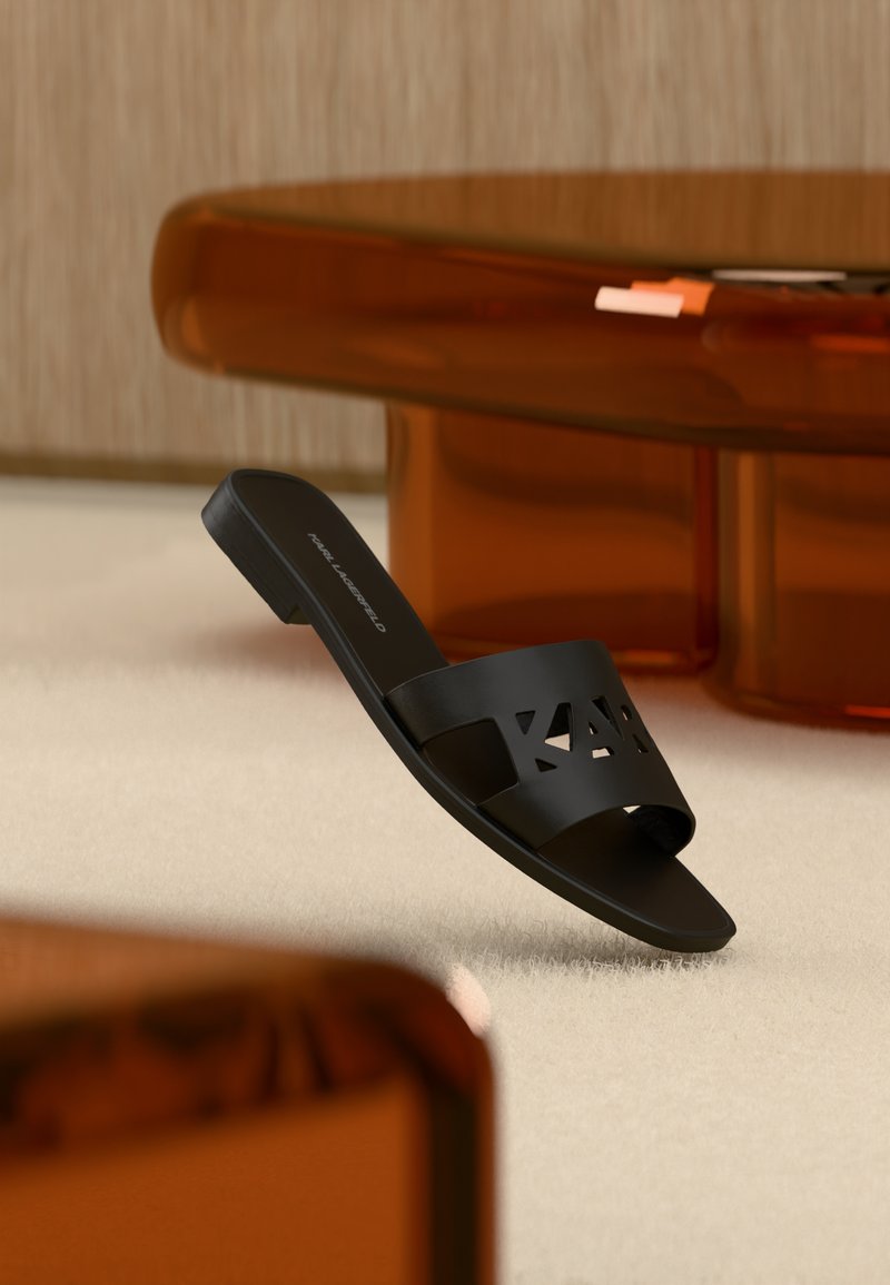 Μαύρο σανδάλι με φάσα και λογότυπο με περικοπή, πάνω σε ανοιχτό χαλί σε δωμάτιο με διάφανα έπιπλα κεχριμπαρένιου χρώματος.